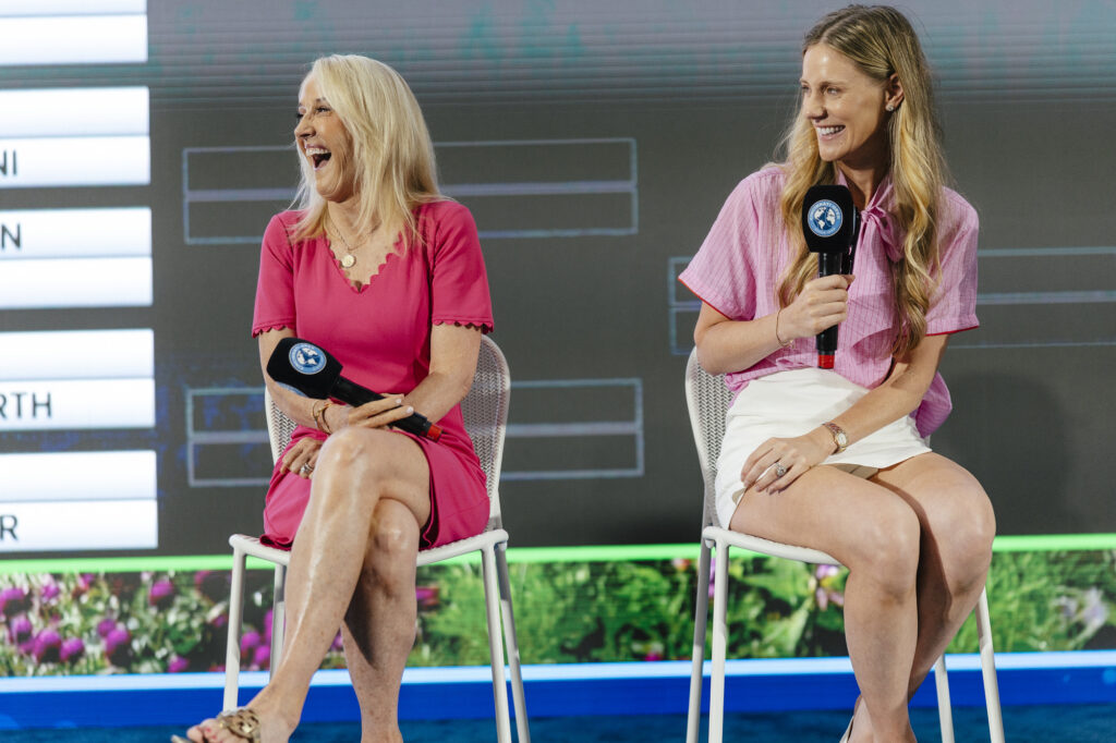 Two women sit on stools on a stage holding branded microphones during a panel discussion, one laughing mid-conversation.