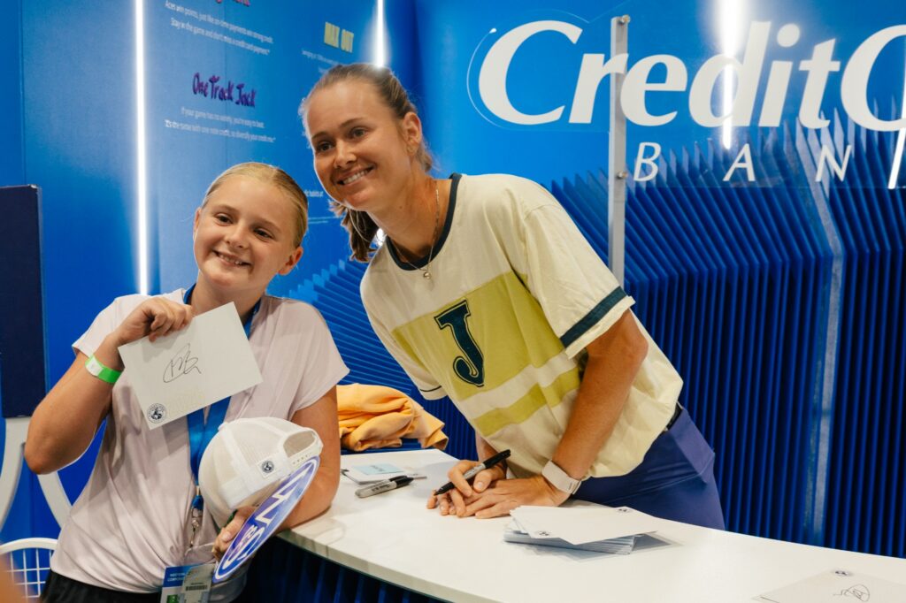 A young fan smiles beside a player at a signing table, holding an autographed card