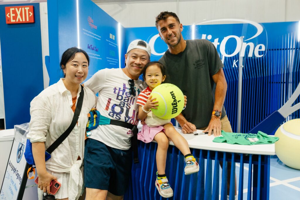 A family poses with a player at a Credit One Bank booth, a child holding an oversized tennis ball.