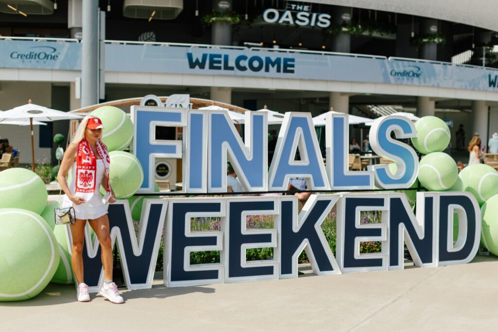 A person poses beside a giant Finals Weekend sign made of oversized letters and tennis balls.