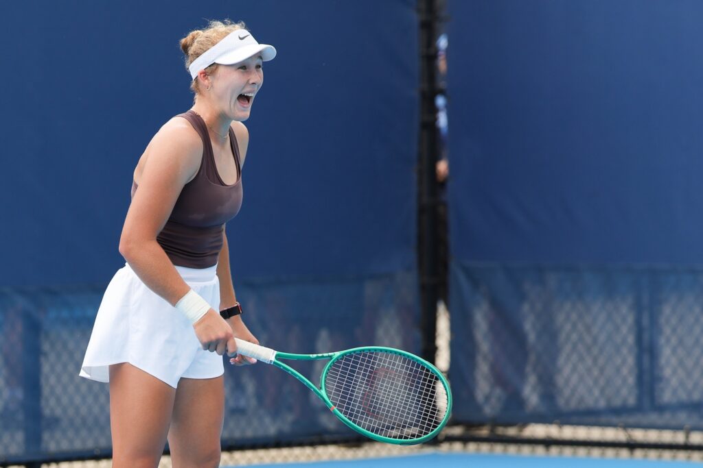 Mirra Andreeva smiles and shouts on the court while wearing a white tennis skirt, white visor and brown tank top