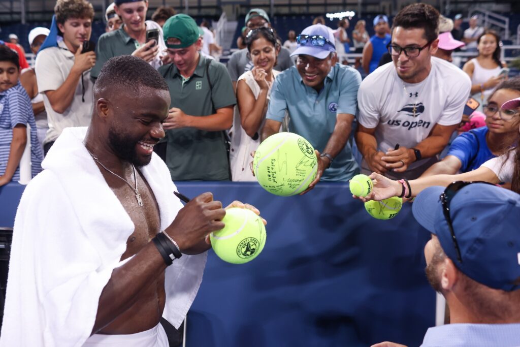 Frances Tiafoe signs oversized tennis balls for fans crowded along the barrier after a night match, a white towel draped over his shoulders and a marker in his hand.