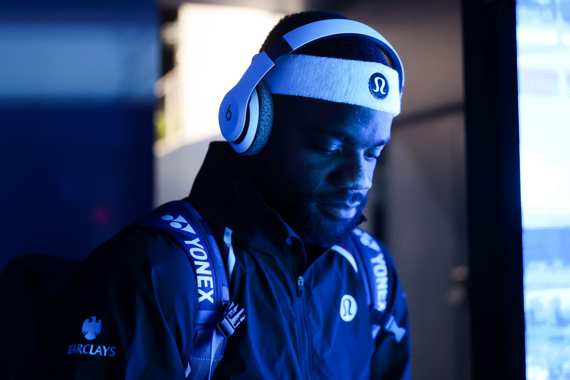 Frances Tiafoe waits in the players' tunnel before a match, lit in deep blue, wearing white over-ear headphones and a headband with his racket bag on his shoulder, eyes down.