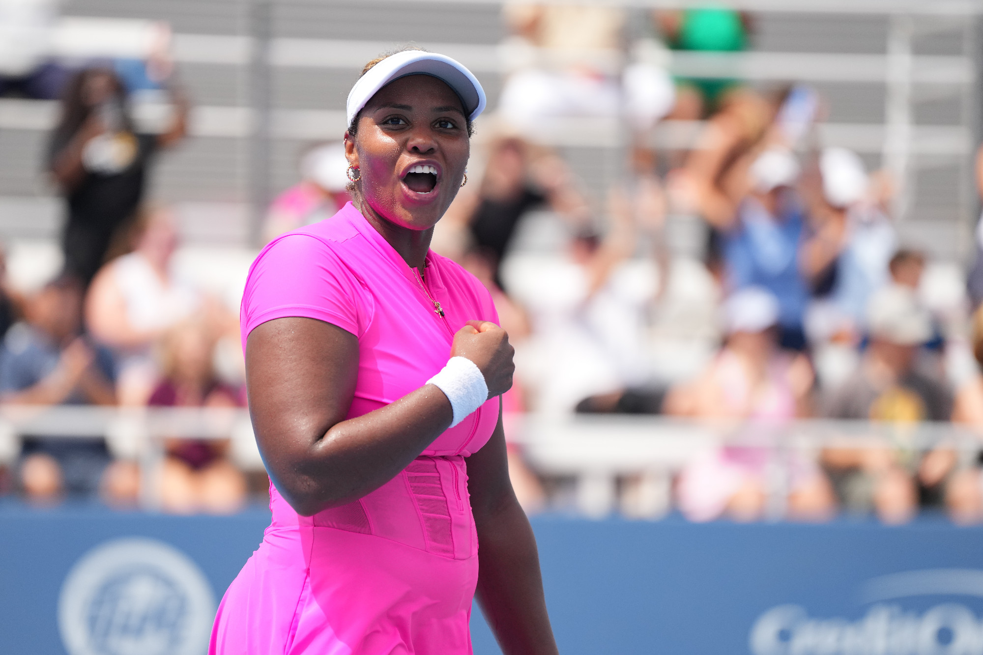 A female tennis player in a bright magenta short-sleeved tennis outfit and white visor pumps her fist and shouts in celebration on a sun-drenched outdoor court. She wears a white wristband and hoop earrings. A packed stadium of blurred spectators fills the background, and a blue courtside barrier with partial sponsor lettering is visible at the bottom of the frame.