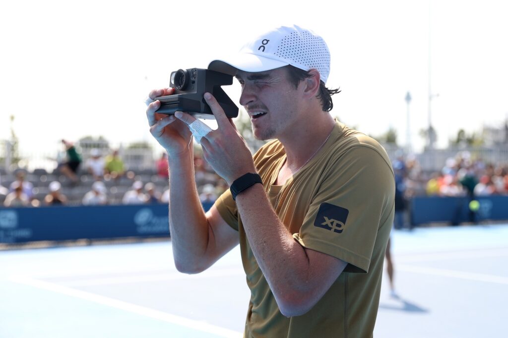 Fonseca uses a polaroid camera whilewearing a brown shirt and white cap