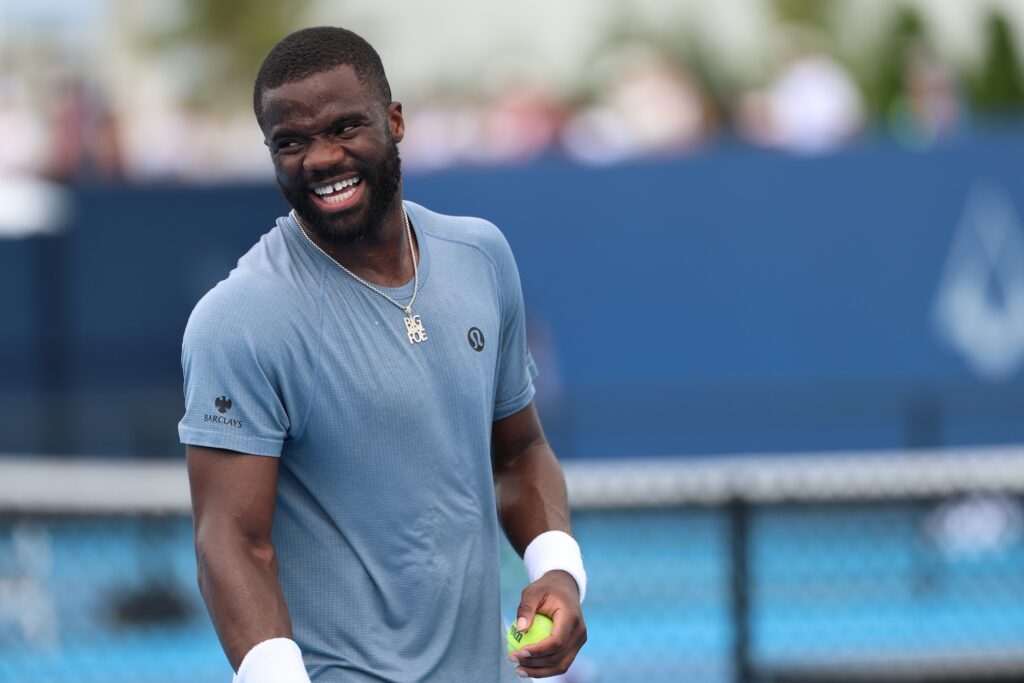 Frances Tiafoe laughs during a practice session at the Cincinnati Open, holding a tennis ball and wearing a blue-grey shirt and a chain necklace.