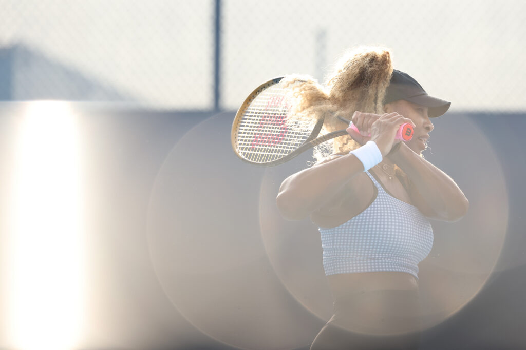 A player practises a two-handed backhand in a checked crop top, backlit by low sun with a lens flare across the frame.