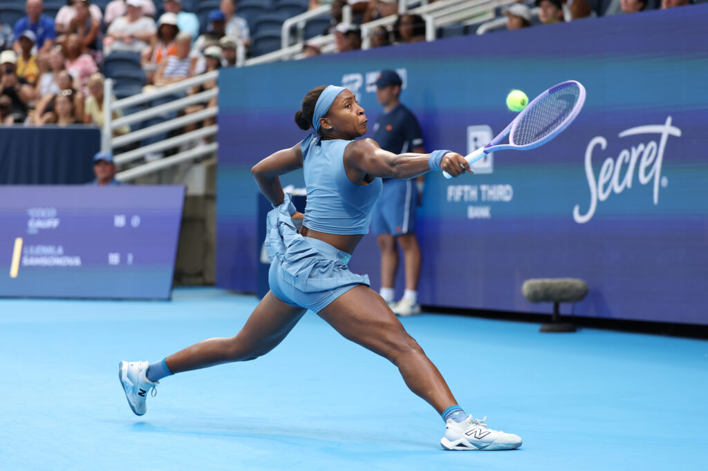 A player runs down a forehand in a pale blue crop top and skirt, the ball just ahead of her racket and a Secret sponsor board behind.