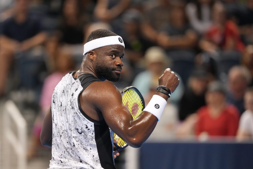 Frances Tiafoe looks back over his shoulder and clenches his fist after winning a point, wearing a white headband and a black and white splatter print tank top.