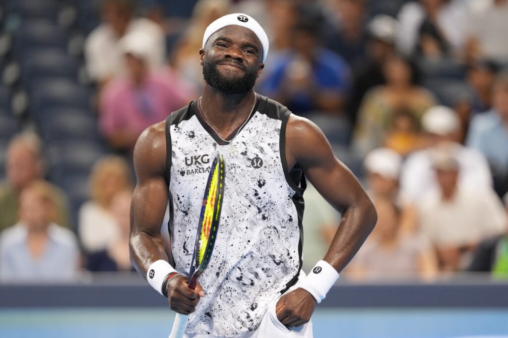 Frances Tiafoe grimaces and pulls at his shorts between points, racket in hand, the crowd blurred behind him.