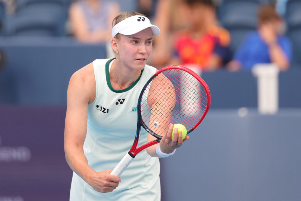Elena Rybakina in white tennis attire and visor prepares to serve, holding a tennis ball and racquet on a tennis court.