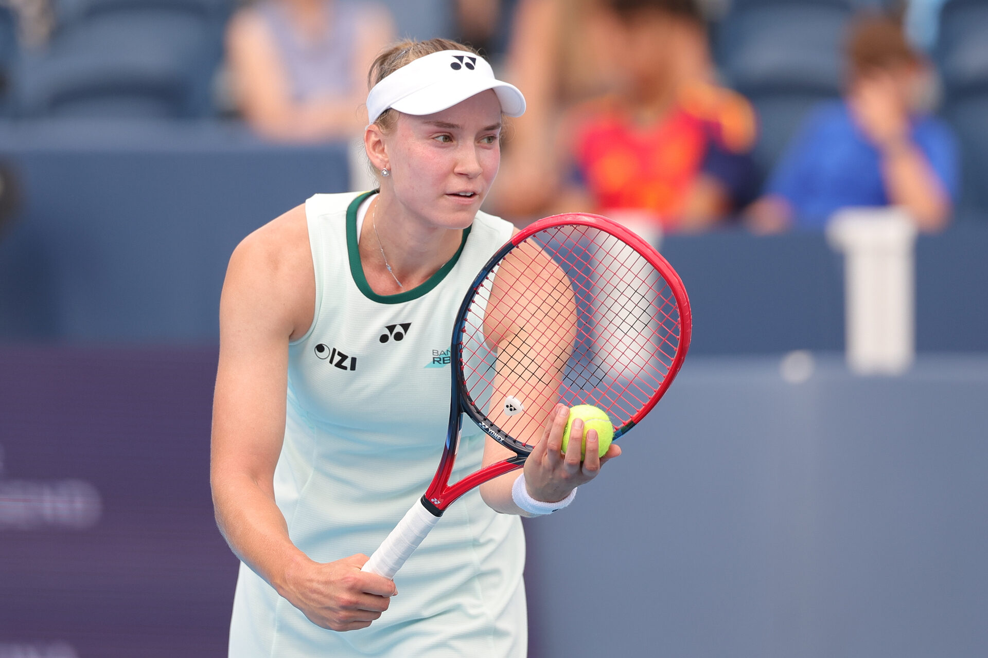 Elena Rybakina in white tennis attire and visor prepares to serve, holding a tennis ball and racquet on a tennis court.