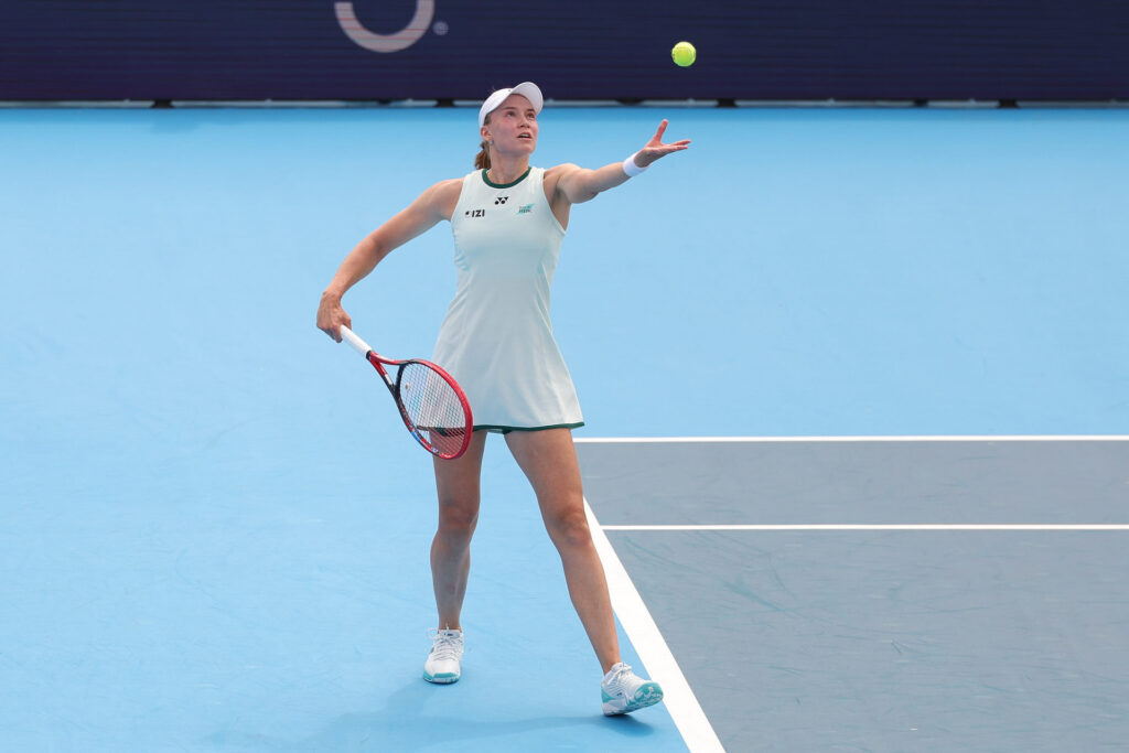 Elena Rybakina is preparing to serve on a blue tennis court, focused on the ball tossed in the air. She is wearing a white sports dress, white visor, and white tennis shoes.