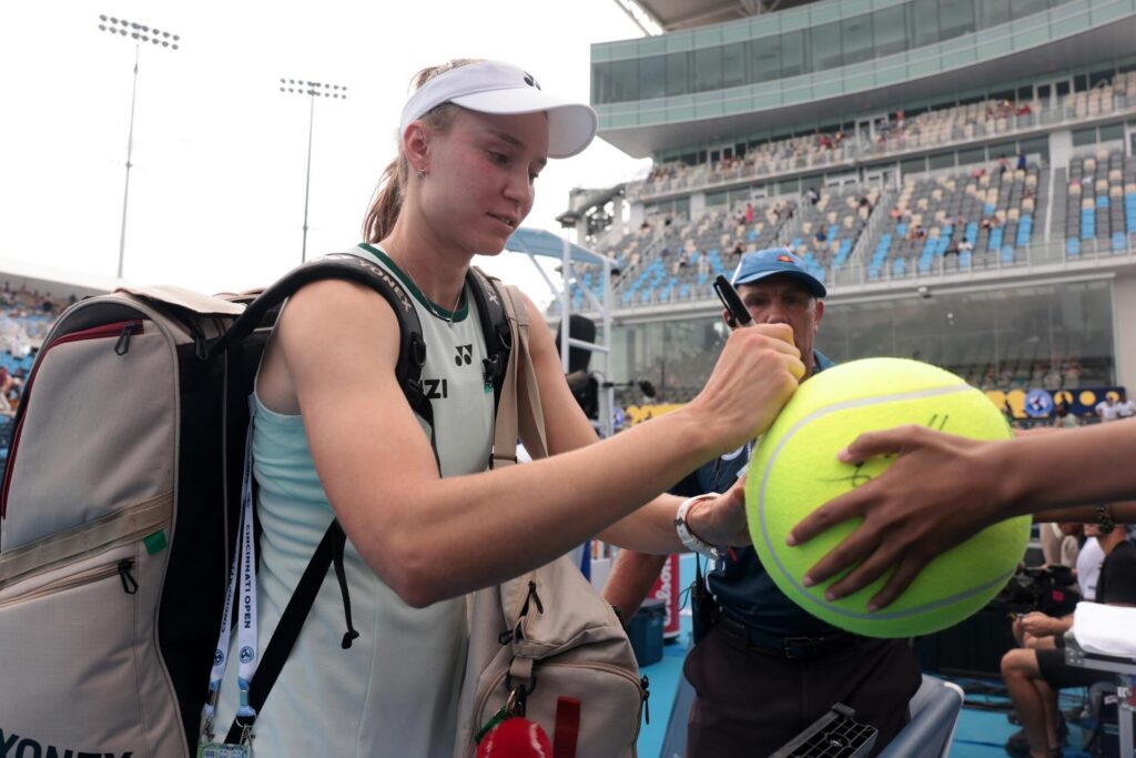 Elena Rybakina is signing a giant tennis ball for a fan at a tennis stadium. She is wearing a visor and carrying multiple tennis bags.