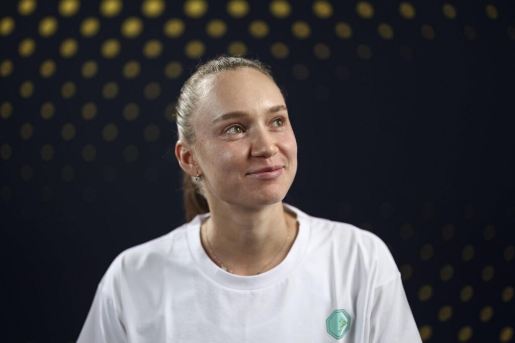 Elena Rybakina is wearing a white t-shirt and looking slightly up with a subtle smile, against a dark background with a pattern of blurred yellow dots.