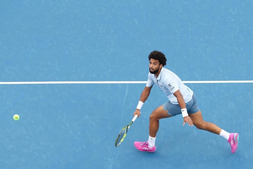 Arthur Fils lunges on a blue tennis court, focused on hitting a tennis ball with his racket. He is wearing a light blue shirt, gray shorts, white wristbands, and bright pink shoes.