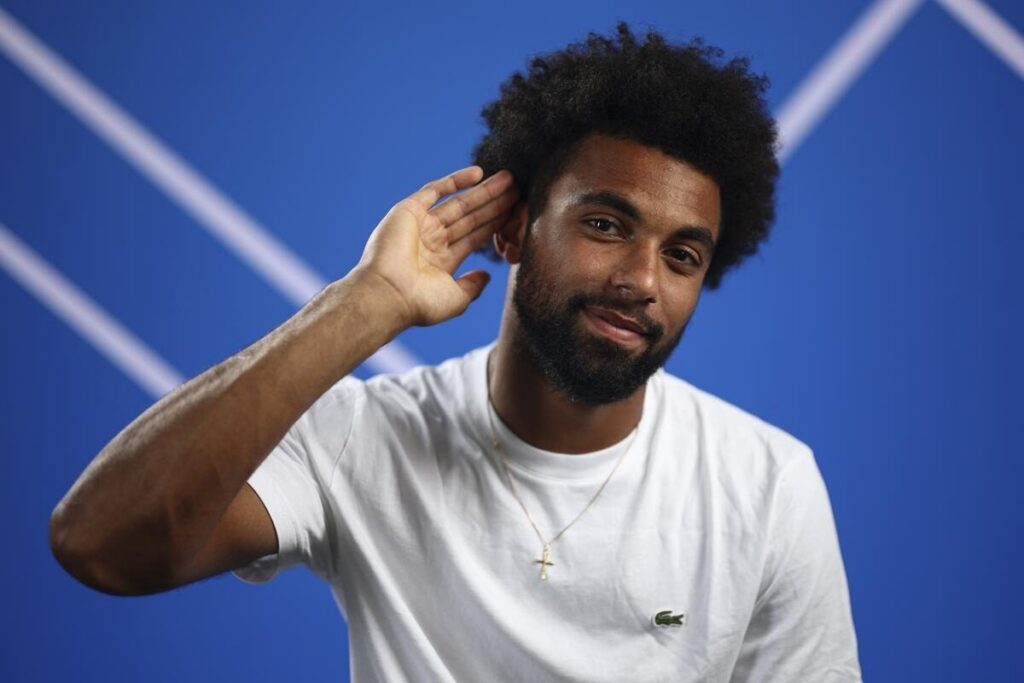 Arthur Fils wearing a white Lacoste t-shirt and a gold cross necklace, holding his right hand to his ear with a slight smile, set against a blue background with white diagonal lines.