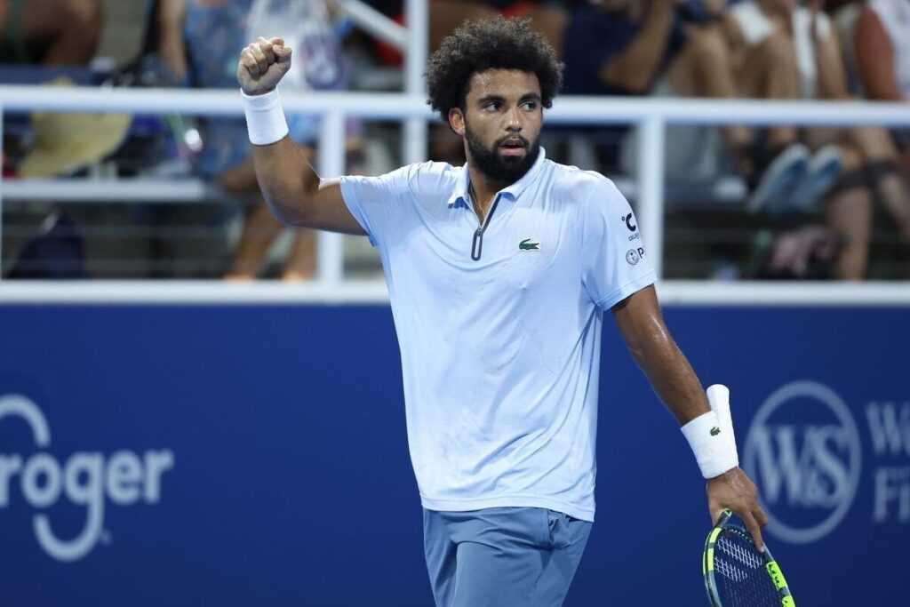 Arthur Fils raises his fist in celebration on a tennis court, holding a racket in his other hand and wearing a light blue shirt and wristbands.