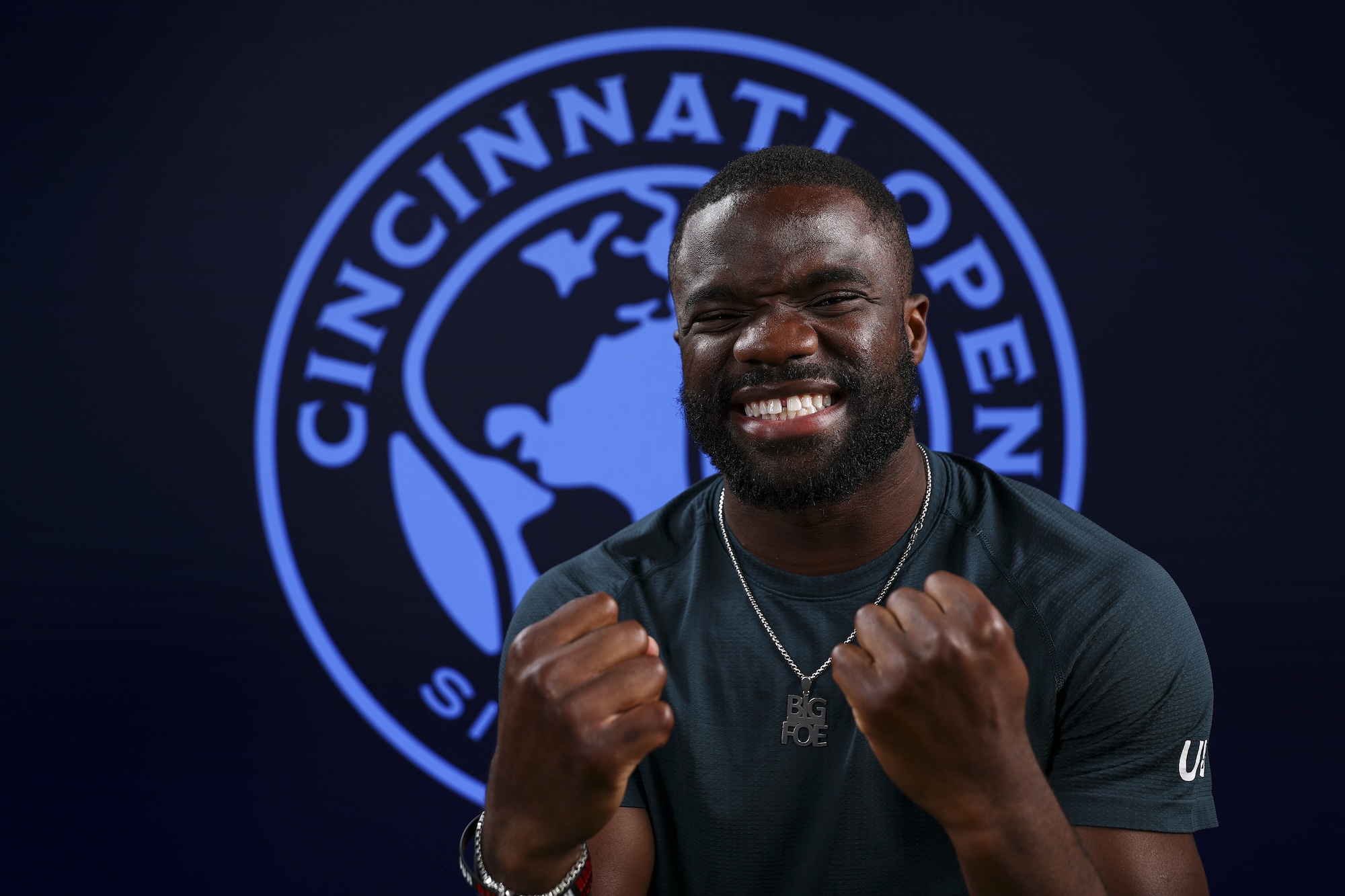 Studio portrait of Frances Tiafoe grinning with both fists raised in front of a large illuminated Cincinnati Open crest, wearing a dark t-shirt and a Big Foe pendant.