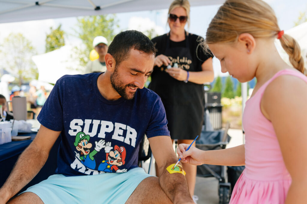 A bearded man in a navy blue Super Mario Bros. t-shirt and light blue shorts sits in a chair under a white event tent, smiling as he watches a young girl paint a small design on his arm. The girl, wearing a pink tank top and skirt, holds a thin paintbrush over a small yellow paint palette. A woman in a black t-shirt and sunglasses stands in the background.