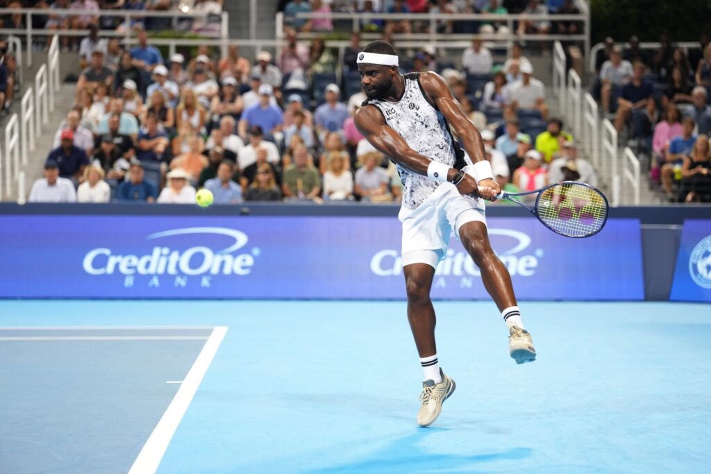A tennis player in mid-swing hitting a backhand shot on a blue hard court, with a crowd watching in the background. The player is wearing a white headband, sleeveless shirt, and shorts.