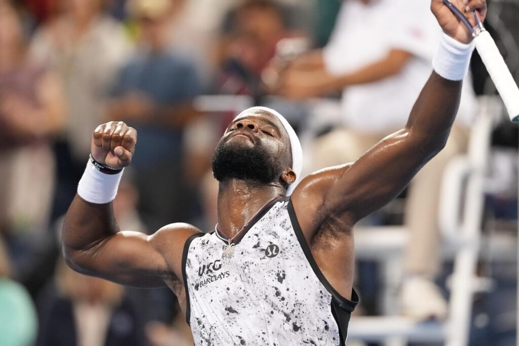 A tennis player wearing a white and black speckled shirt and headband raises his arms in celebration, with his eyes closed and a determined expression. He is sweating and holds a tennis racket in his right hand.