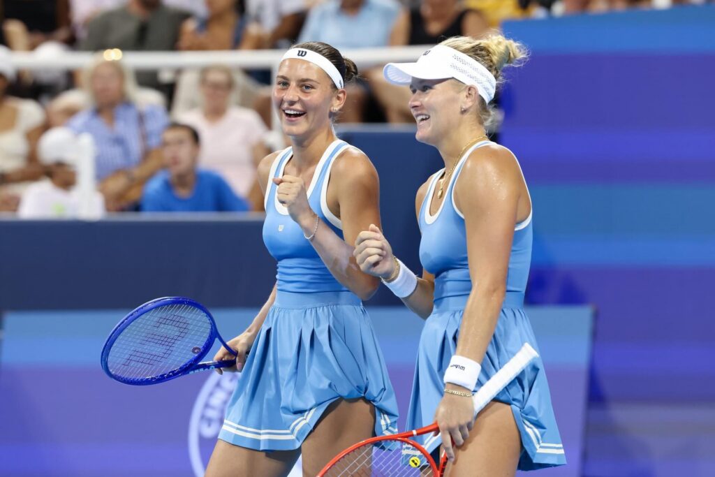 Two female tennis players in matching blue outfits smiling and celebrating on the court, each holding a tennis racket.