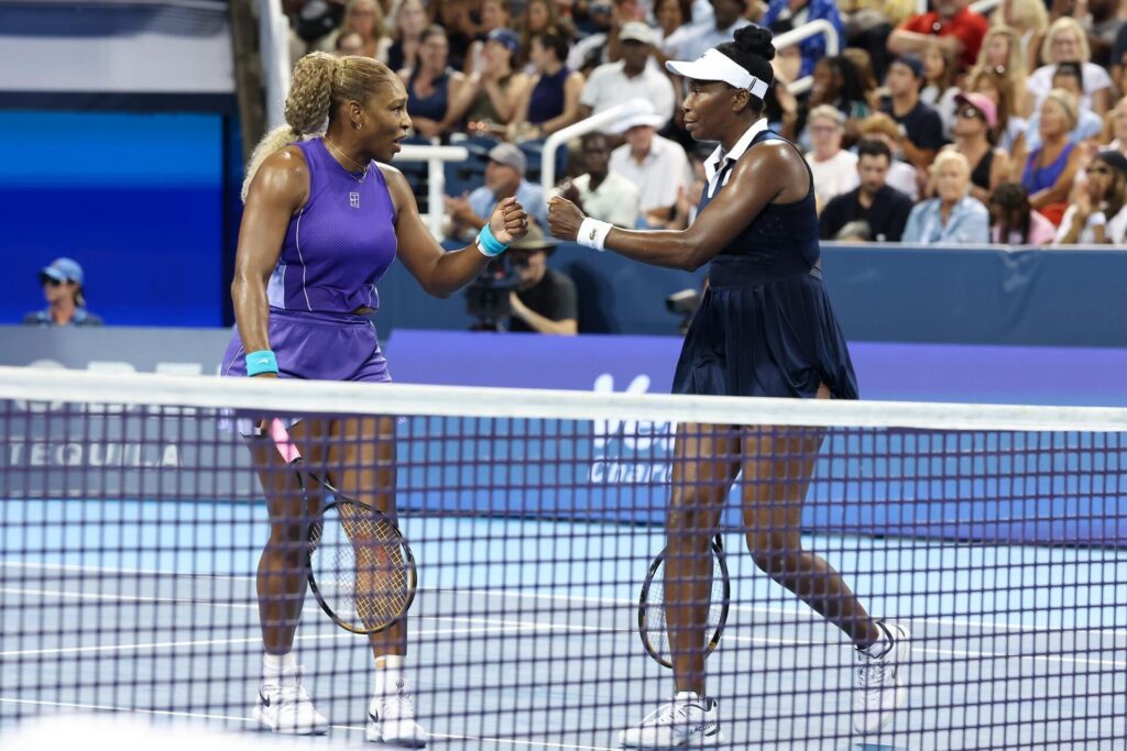 Two female tennis players fist bump each other across a tennis net during a match, with an audience watching in the background.