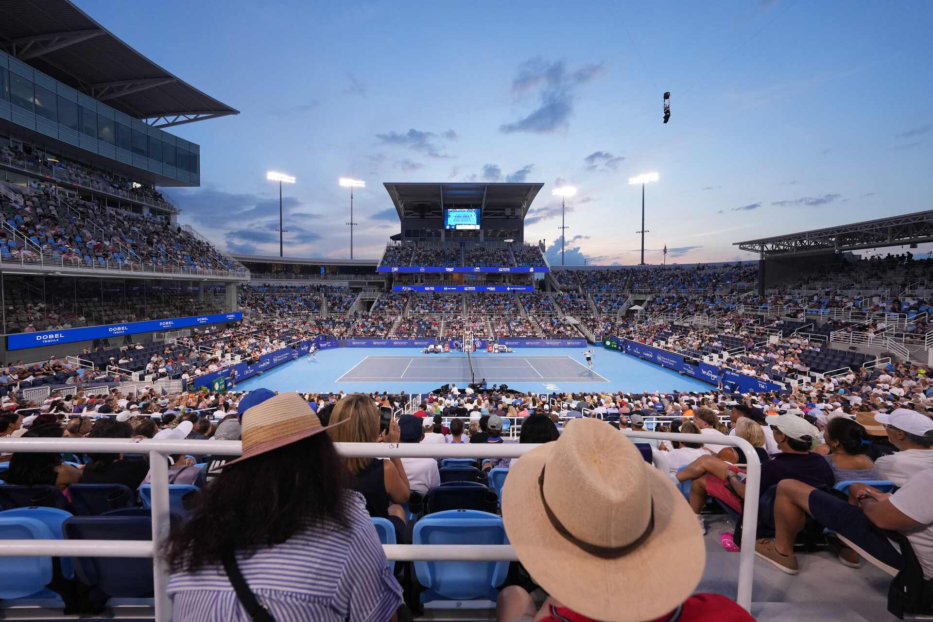 Large outdoor tennis stadium filled with spectators watching a match during sunset, with players visible on the court and stadium lights on.