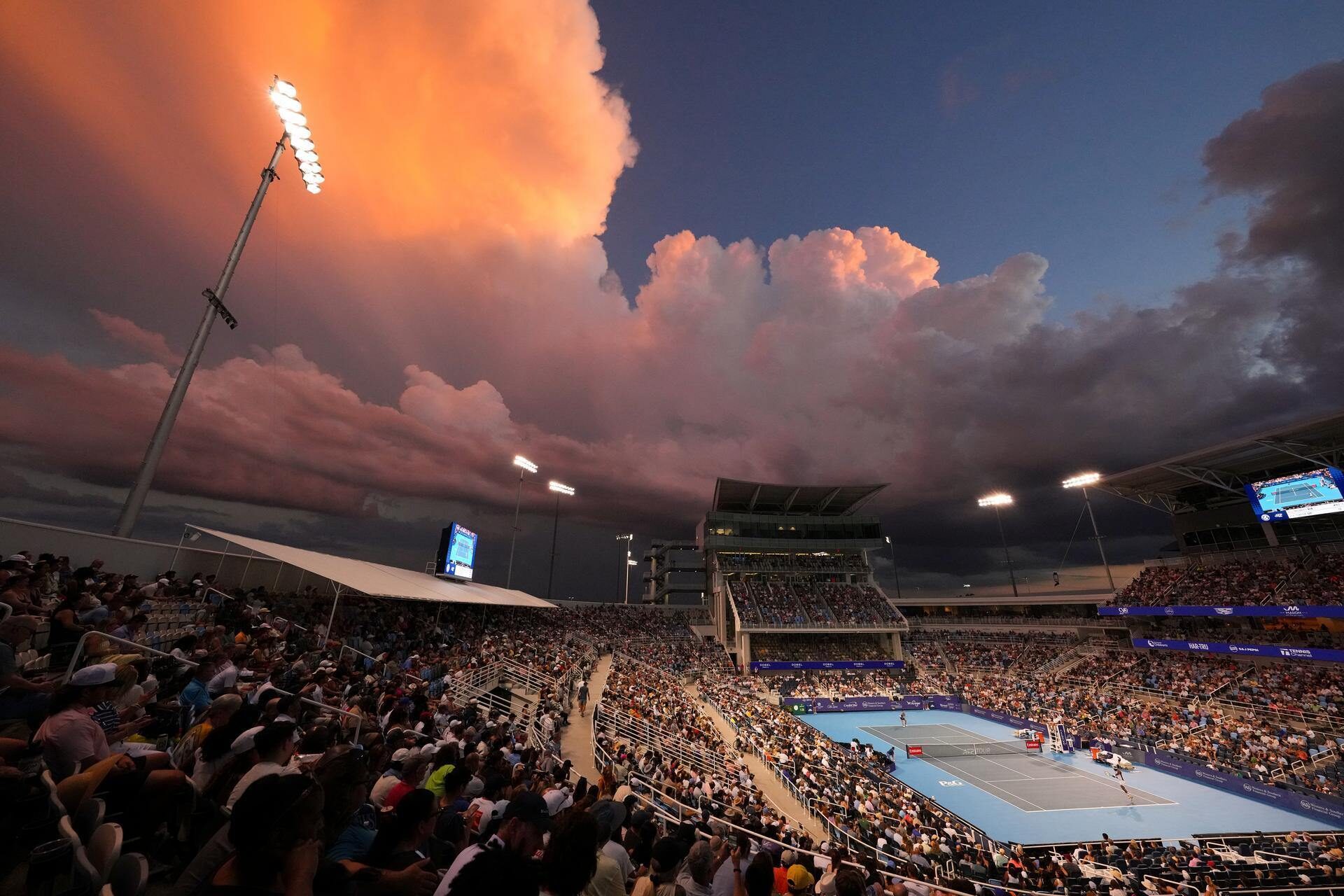 A large crowd watches a tennis match at an outdoor stadium during sunset, with dramatic orange and purple clouds in the sky. Stadium lights illuminate the court and spectators.
