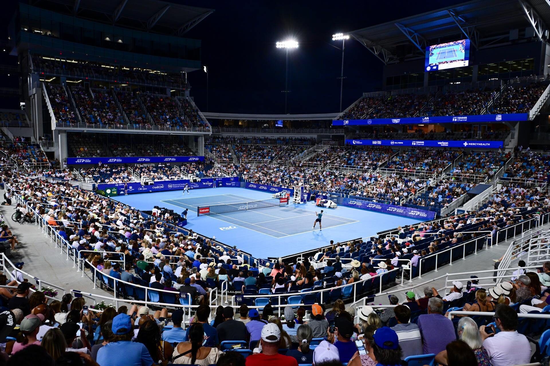 A night tennis match takes place on a blue court inside a large, well-lit stadium filled with spectators. Two players are actively engaged in the game.