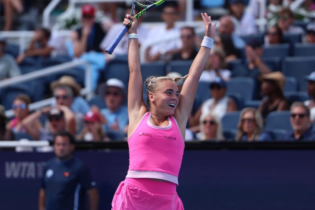 Sara Bejlek in a pink outfit raises her arms and racket in celebration on the court, with spectators blurred in the background.