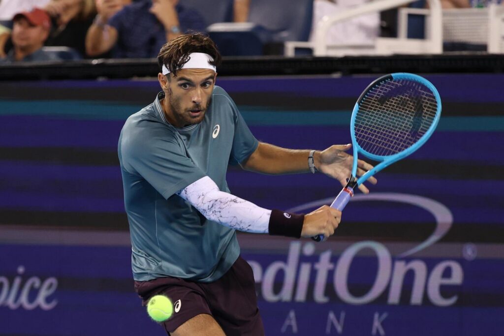 A male tennis player wearing a blue shirt and white headband prepares to hit a backhand shot with a blue racket. A tennis ball is approaching him, and spectators are visible in the background.