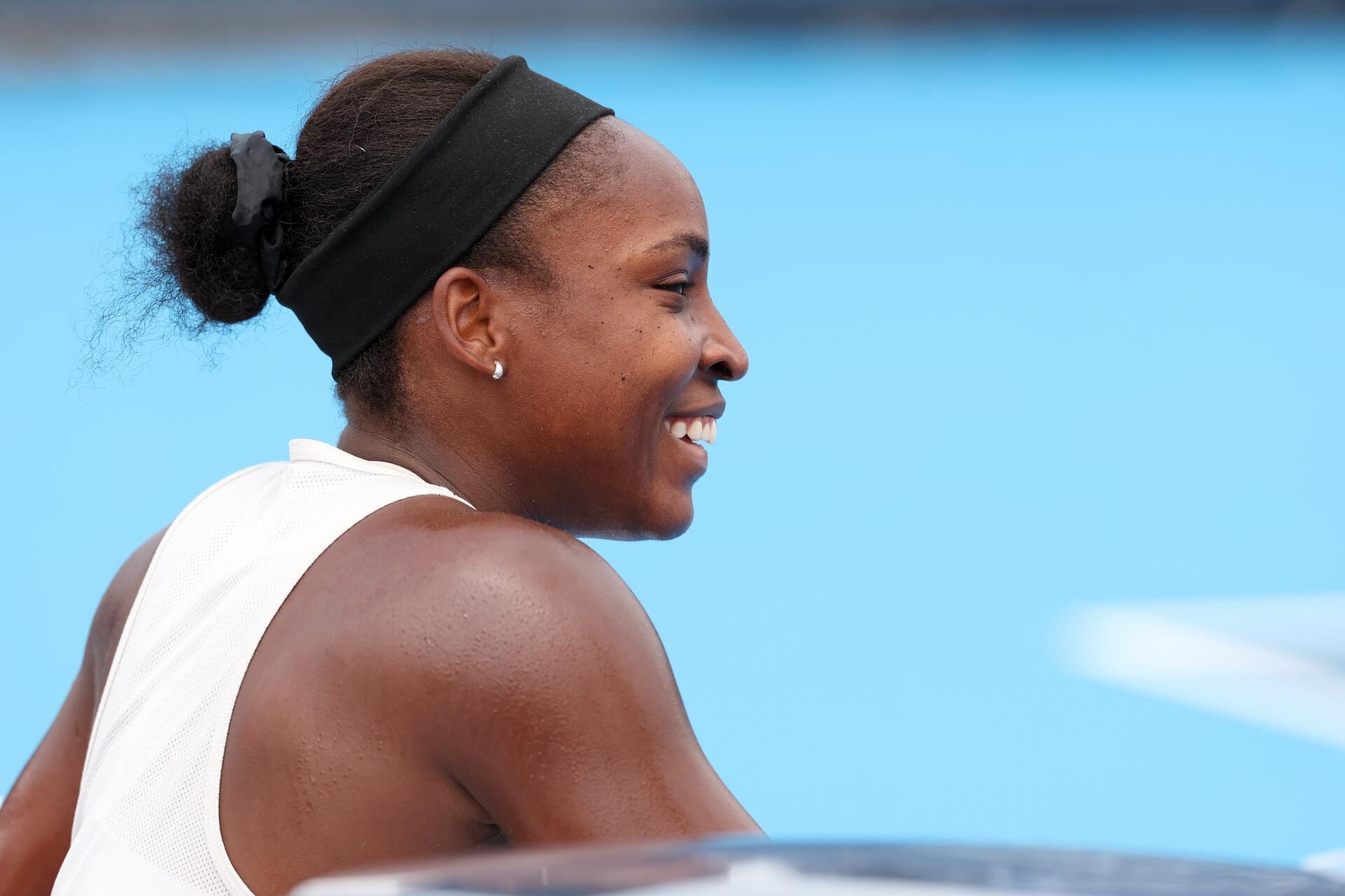 Coco Gauff in a white sleeveless top and black headband, smiling and looking to the right against a bright blue background.