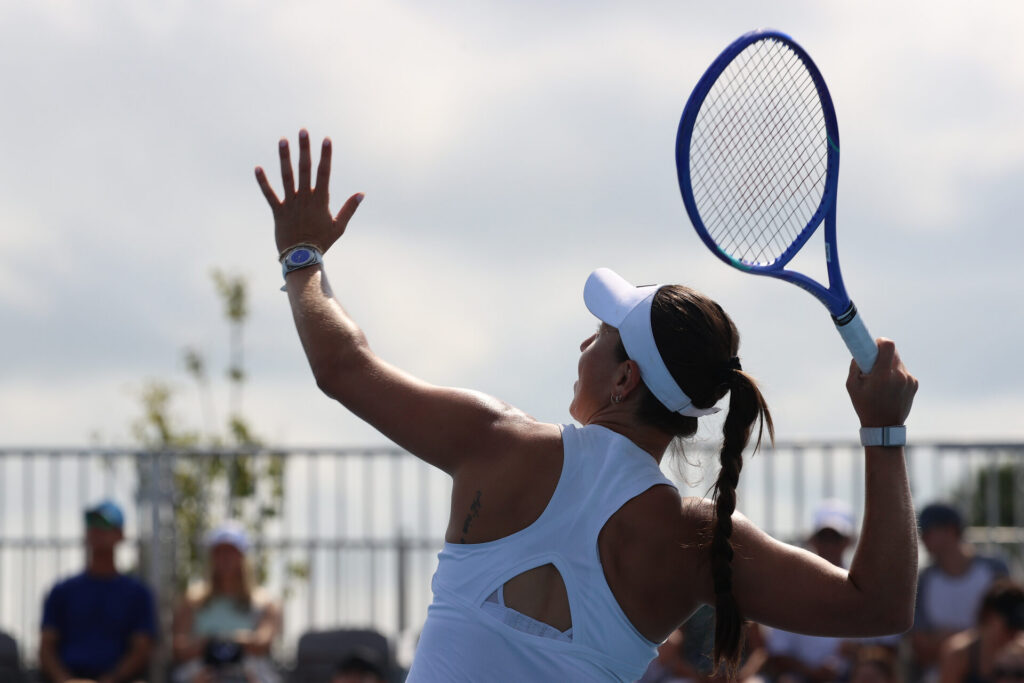 Pegula prepares to serve a tennis ball, holding a tennis racket behind her head with her right hand while stretching her left hand upward.