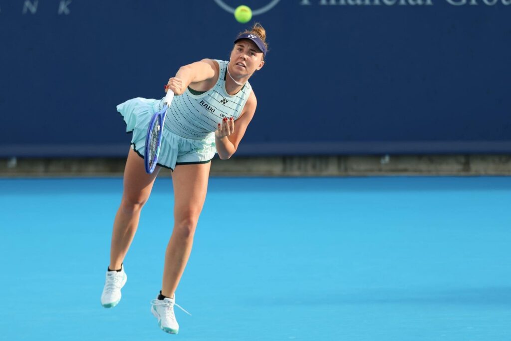 Linda Noskova serves the tennis ball intensely on a bright blue court wearing a light blue dress and visor. She is focused, with her racket extended forward and one foot slightly off the ground.