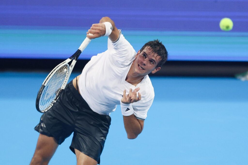 Rafael Jodar in mid-serve during a tennis match on a blue court, wearing a white shirt and black shorts.