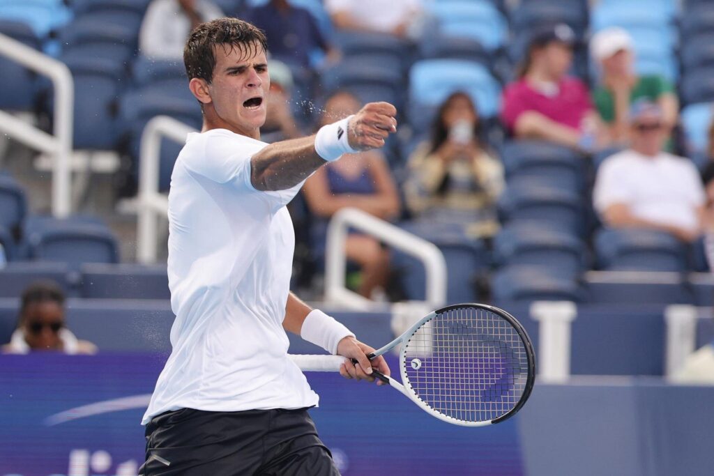 Rafael Jodar is captured mid-action during a tennis match, wearing a white shirt and black shorts, with an intense expression and a tennis racket in his hand. Spectators are visible in the blurred background.