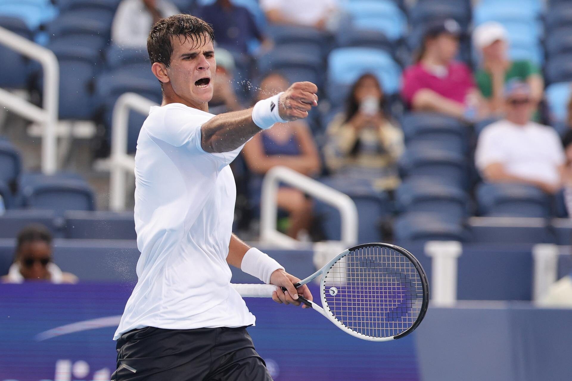 Rafael Jodar is captured mid-action during a tennis match, wearing a white shirt and black shorts, with an intense expression and a tennis racket in his hand. Spectators are visible in the blurred background.