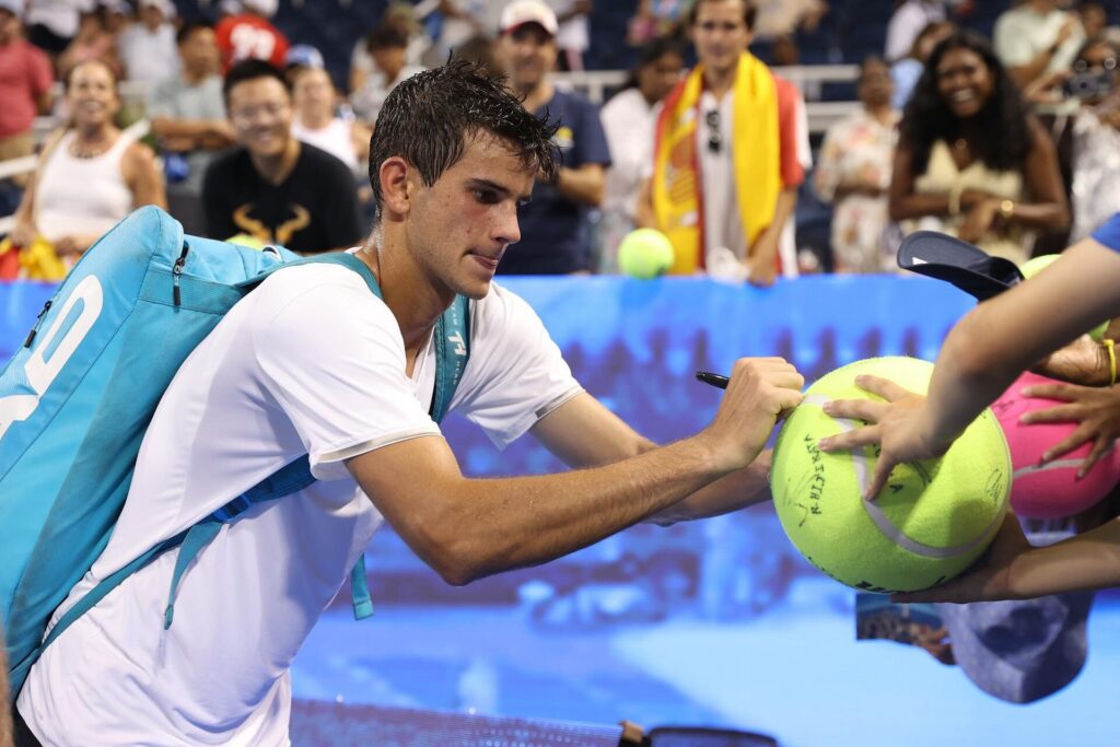 Rafael Jodar, wearing a white shirt and a blue backpack, signs a large tennis ball for fans after a match.