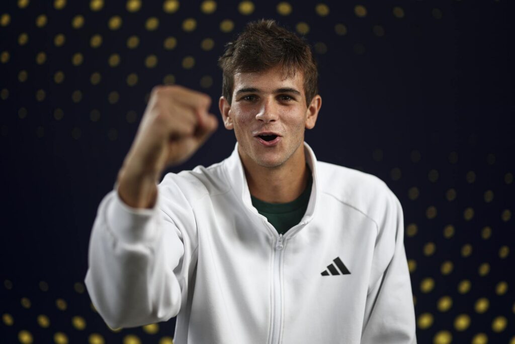 Rafael Jodar wearing a white Adidas jacket with a raised fist and an excited expression, against a dark background with yellow dots.